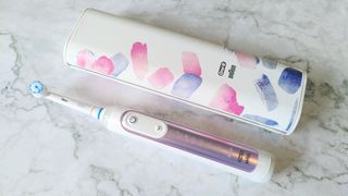 En vit Oral-B Genius X eltandborste med rosa detaljer ligger på ett marmormönstrat bord bredvid sitt tillhörande vita fodral med rosa och blå detaljer.