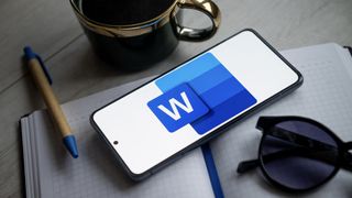 Microsoft Word logo on mobile sitting on notepad next to mug, pen, and sunglasses