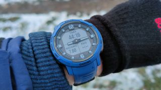 The Garmin Instinct Crossover smartwatch pictured in blue on someone's arm with a snowy, grass background.