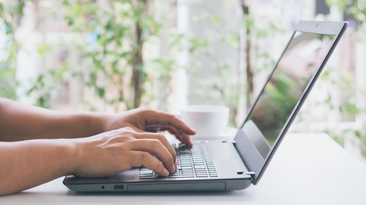 Primer plano de las manos de una persona trabajando en el teclado de un portátil y el portátil descansando sobre una mesa en una habitación iluminada por el día.