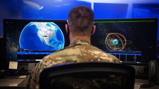 a person in camouflage sits at a computer screen looking at an animation of several rings around Earth denoting spacecraft orbits 