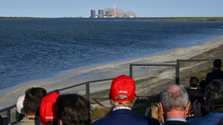 a crowd of people, including a man in a red baseball cap, watch a rocket launch from across a stretch of water