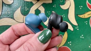 A JBL Live Beam 3 earbuds and a Tour Pro 3 earbud, held in a hand, to show the relative depth