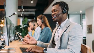 A side on view of a man in a call center happily talking to a customer on the phone while looking at his compuer screen and typing on a keyboard. There are three colleagues out of focus sitting alongside him