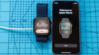 Close up of an Apple Watch 10 screen showing "Hello" next to an iPhone with the same message. Both are sitting on a light blue mat