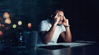 Software developer looking stressed at desk in a dark room with monitor light illuminating face.