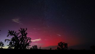 a long white tail of a meteor streaks across the sky which is colored a deep red from the auroras.