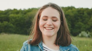 Image depics a young women with dimples on both cheeks, smiling in a field