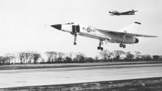 a jet plane landing on a runway accompanied by another plane behind it, in mid-air. the image is in black and white due to age. bare trees are visible in the background