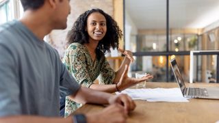 A young, female employee talking in a friendly manner with a male colleague. Both represent Gen Z in the workplace and are sat at a desk in a modern office.