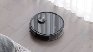 En svart Ecovacs Deebot Ozmo T8 AIVI städar ett mörkt trägolv bredvid en vit gardin.