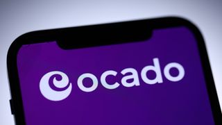 The Ocado logo in white, against a purple background shown on a phone. Decorative: The phone is in extreme-close-up to the camera and the Ocado logo is the word "Ocado" in white with a stylized swirl to the left.