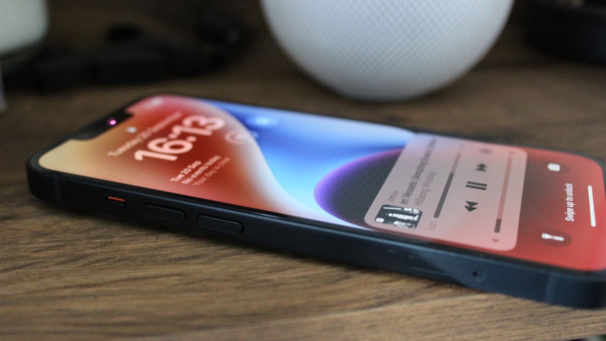 The iPhone 14 pictured on a wooden table showing the homescreen and a music track playing.