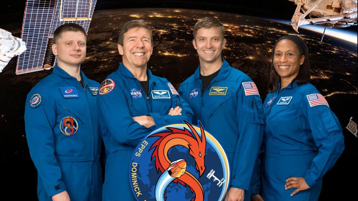 three white men and a black woman stand smiling wearing blue astronaut flight suits. the circular graphic of the crew&#039;s mission patch, with an orange dragon spiralled into an 8, is in front of the group. a view of Earth at night, as seen from the space station is their backdrop.