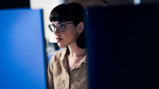 Female cyber security worker with glasses sitting at a workstation in an office environment.