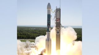 Rocket launching from pad, cloudy plumes billow from beneath the rocket as it takes off. The sky is clear and blue.