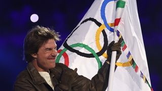 Tom Cruise, dressed in a brown leather jacket, waves the Olympic flag at the Closing Ceremony of the Paris 2024 Games.