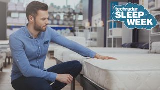 A man in a blue shirt and black jeans lays his hand on a mattress that he wants to buy