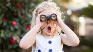 Little girl has a shocked expression while using one of the best binoculars for kids with a foliage backdrop