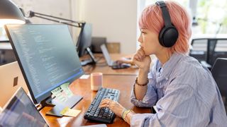 A female software engineer working at a desk in an office, with code on her screen. Decorative: She is wearing a blue dress shirt and has short, pink hair.