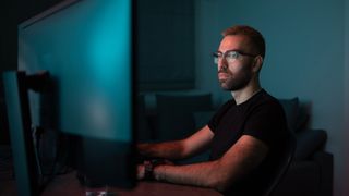 A photo of a software developer sat at a desk from the chest up, with the both of their monitors turned away from the camera to represent shadow IT. Their face is lit by the screens in an otherwise dark room.