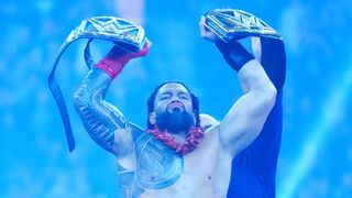 WWE wrestler Roman Reigns stands in the ring triumphantly holding two belts aloft.