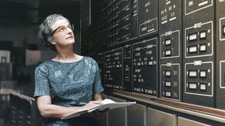 Nancy Grace Roman standing holding some papers and a pen while looking at the wall on the right.