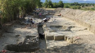 Several workers excavate an ancient Roman city by a field with trees.