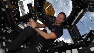 an astronaut in a blue shirt floats in a space station module surrounded by windows. earth can be seen through the windows