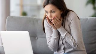 A shocked girl sitting on sofa at home looking on laptop screen