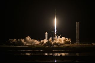 a white rocket lifts off at night above a plume of fire and smoke