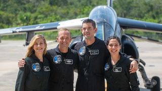 Four people in dark jumpsuits stand in a row in front of a jet.