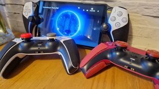 Two DualSense controllers and a PlayStation Portal with thumbsticks and grips attached on a wooden surface with a white brick background