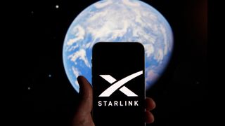 In the foreground is a hand holding a phone with the Starlink logo on the screen behind it in the background of the image is planet Earth.