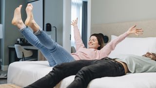 A man lies down on a bed while a woman jumps onto the bed next to him