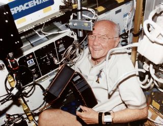 An older light-skinned man with grey hair on the sides of his balding head, is crouched down near a wall of electronics racks. Cables and cords are wrapped and plugged in throughout the space. He is smiling, and holding near his leg to black components.