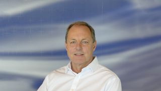 Dr Dave Smith, the new UK National Technology Adviser, sat in a white shirt against a background of solid blue broken up with clouds and white cross marks.