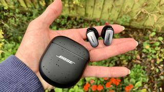 Bose QuietComfort Ultra oordopjes in een hand met de case, boven een bloembed.