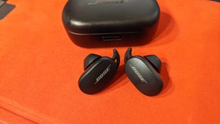 Et par svarte Bose QuietComfort Earbuds ved siden av det tilhørende ladeetuiet, liggende på en oransje flate.