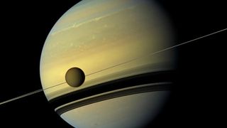 saturn hangs huge, the shadows of its rings striping its lower half. a moon hangs before the giant's body, sliced by the thin line of rings behind.