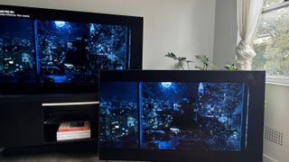 The LG G4 OLED TV on a TV bench, with LG G3 OLED next to it on the floor, and both showing the same scene of a man being thrown out of a glass window, in the movie Watchmen