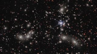 A crowded galaxy field on a black background, with one large star dominating the image just right of center. Three areas are concentrated with larger white hazy blobs on the left, lower right, and upper right above the single star. Scattered between these areas are many smaller sources of light; some also have a hazy white glow, while many other are red or orange.