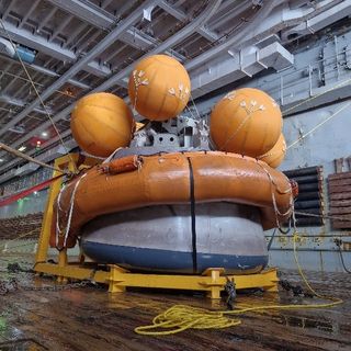 a large metal capsule sits on a yellow metal frame on a wooden deck with an orange float ring around the bottom and six large orange floats near the top.