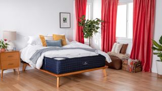 DreamCloud Mattress sales, deals and discounts: The DreamCloud Mattress shown on a light wood frame bed with red drapes hanging at the windows