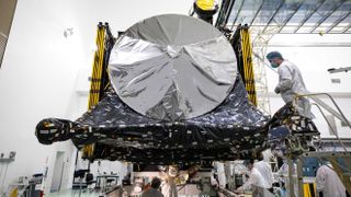 technicians in white coats work on a shiny silvery spacecraft inside a white-walled room.