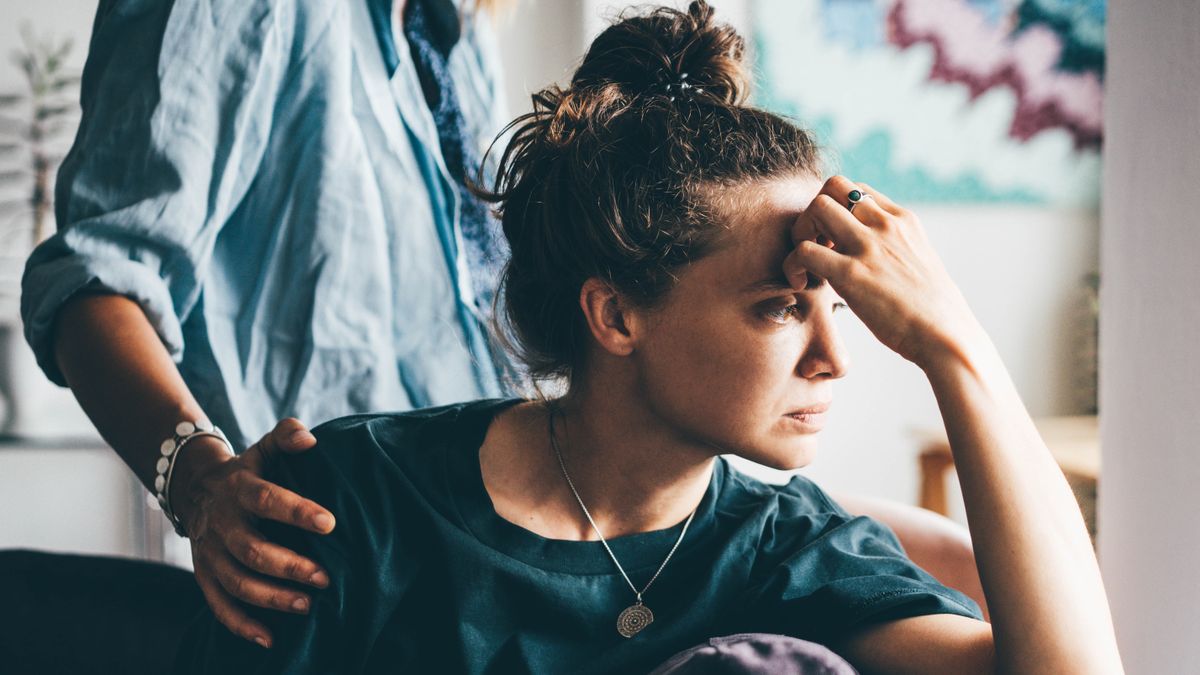 a photo of a woman sitting down with one hand on her head, as if frustrated; a person standing behind her has a hand on the woman&#039;s shoulder