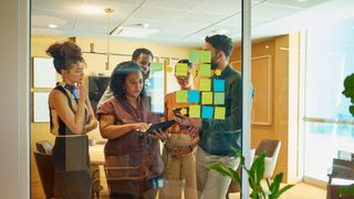 A diverse group of employees look at post-it notes on a large window inside a modern office, while the one in the center of frame holds a tablet.