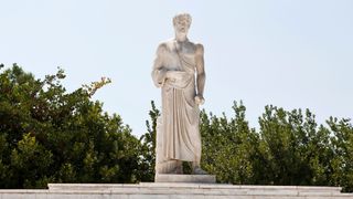 A white marble statue of Hippocrates standing at the top of a set of marble stairs near some trees and bushes