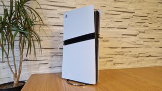 The PS5 Pro in front of a white brick background, on a brown desk, next to a potted plant.