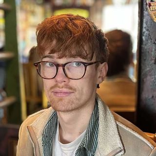 A head and shoulders photo of George Fitzmaurice, with short red hair, glasses and a tan jacket over a stripey green shirt and white t-shirt.